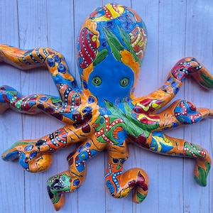 May include: A colorful ceramic octopus wall hanging. The octopus features a blue head, orange tentacles, and is decorated with intricate patterns in red, yellow, green, and blue. The octopus has large, round eyes and is mounted on a light blue wooden surface.