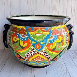May include: A large, round, hand-painted ceramic planter with a vibrant, colorful design. The planter features a black rim and handles, and is decorated with intricate patterns in shades of blue, orange, yellow, and green. A decorative piece for the home or garden.