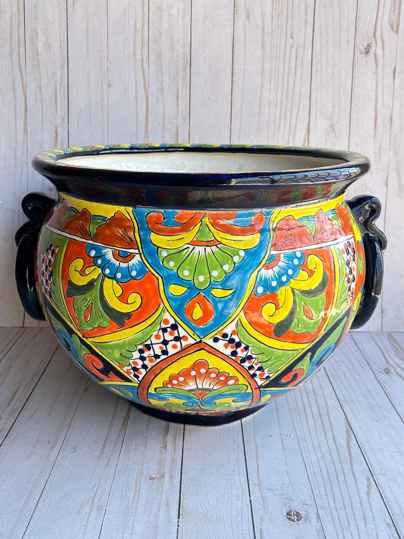 May include: A large, round, hand-painted ceramic planter with a vibrant, colorful design. The planter features a black rim and handles, and is decorated with intricate patterns in shades of blue, orange, yellow, and green. A decorative piece for the home or garden.