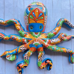 May include: A ceramic octopus wall sculpture featuring a vibrant, multi-colored hand-painted pattern. The octopus has a glossy finish with bright orange, green, blue, and white floral and geometric motifs covering its head and eight curved tentacles.