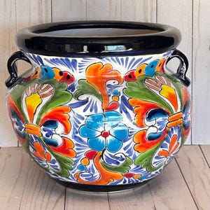 May include: A round ceramic planter featuring a glossy black rim and two side handles. The exterior is decorated with a vibrant, multi-colored floral and scroll pattern in shades of orange, blue, green, yellow, and red against a white background.