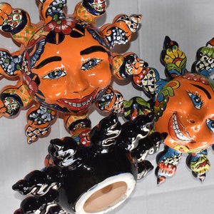 May include: Three ceramic sun sculptures with colorful painted details. The suns have smiling faces with black eyes and red lips. The suns have a black, white, and orange color scheme.