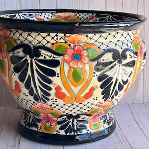 May include: A large, hand-painted ceramic planter with a wide rim and a pedestal base. The planter features a black, white, orange, green, and blue floral design. The design includes stylized flowers and leaf patterns. The planter has two small handles.