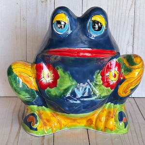May include: A glossy ceramic frog figurine featuring a deep blue body with a bright red mouth. The frog is decorated with hand-painted red flowers, green leaves, and yellow accents on its limbs and eyes. The surface has a smooth, reflective finish.