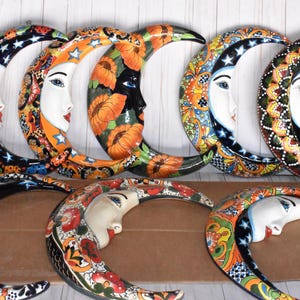 May include: A collection of eight colorful ceramic crescent moon wall hangings. Each moon features a stylized face with intricate floral and geometric patterns in shades of blue, orange, yellow, and black.