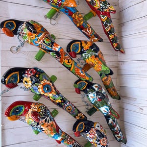 May include: A collection of nine ceramic parrot figurines, each featuring vibrant, hand-painted floral and geometric patterns in colors including red, blue, yellow, orange, and black. Each parrot is attached to a green perch with a metal chain.