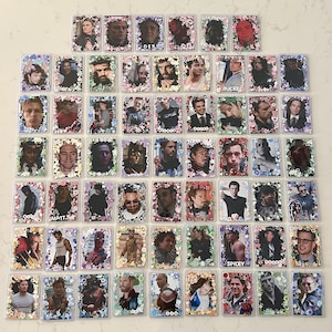May include: A collection of rectangular trading cards featuring various male characters from films, each encased in a clear plastic sleeve and decorated with colourful, small floral and heart-shaped stickers along the borders of the cards.