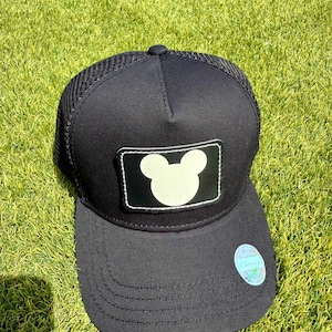 May include: Black baseball cap with a white Mickey Mouse silhouette patch. The cap has a mesh back and a curved brim. A small sticker is on the brim. The cap is sitting on green grass.