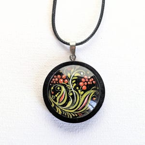 May include: A round wooden pendant necklace with a black cord. The pendant features a colorful floral design with red berries and yellow and green leaves on a black background.