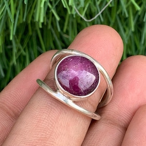 May include: A silver ring with a large, round, pink ruby gemstone set in the center. The ring has a simple, modern design with a thin band that wraps around the stone.