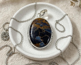 Puede incluir: Un collar con colgante ovalado que presenta una piedra preciosa pietersita pulida con patrones de remolinos azul profundo y marrón dorado, engastada en un marco de metal plateado con borde de cuentas y una cadena de eslabones plateada.