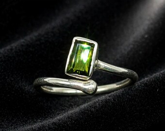 May include: A silver bypass ring featuring a rectangular, faceted green gemstone set in a smooth metal bezel. The band has a polished, rounded finish and an open design, with one end terminating in the gemstone setting and the other in a rounded tip.