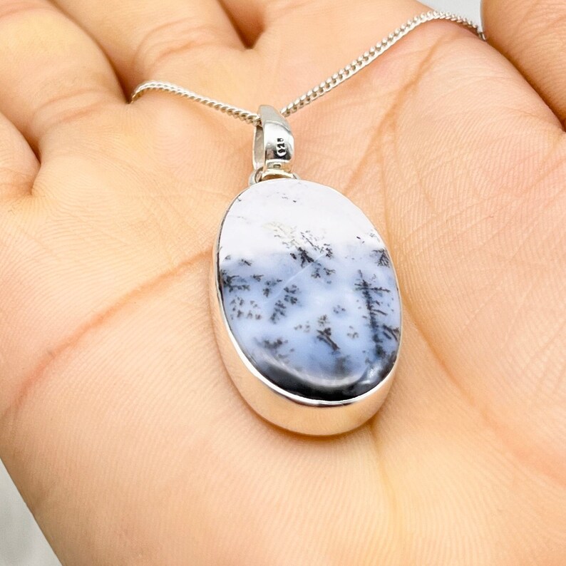May include: Oval pendant necklace with a silver chain. The pendant features a white and grey stone with black dendritic inclusions. The stone is set in a silver bezel. The necklace is displayed on a hand.