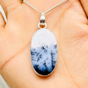 May include: Oval pendant necklace with a dendritic agate stone. The stone has a landscape pattern in white, grey, and black, set in a silver-coloured metal frame. The pendant hangs from a delicate silver chain.