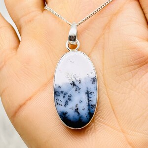 May include: Oval pendant necklace with a dendritic agate stone. The stone features a landscape design in shades of white, grey, and black, set in a silver-tone metal frame. The pendant hangs from a delicate silver chain.
