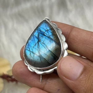 May include: A silver pendant featuring a teardrop-shaped blue labradorite gemstone. The pendant has a detailed silver border and a pin on the back.