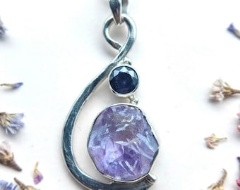 May include: A silver pendant featuring a large, irregular, raw purple amethyst stone at the base and a smaller, round, faceted dark blue gemstone set above it. The silver frame has a curved, open design that loops at the top to hold the chain.