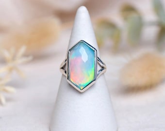 Puede incluir: Un anillo de plata con una gran piedra preciosa de ópalo facetada de forma hexagonal. La piedra muestra destellos iridiscentes de colores azul, verde y rosa. La banda del anillo es de plata pulida y tiene un diseño dividido en los lados.