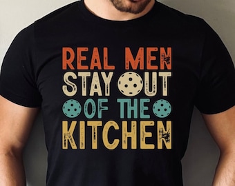 May include: A black t-shirt with the text "Real Men Stay Out Of The Kitchen" in a vintage style font. The text is surrounded by three images of pickleball paddles.