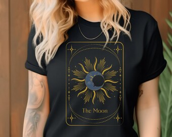 May include: Black t-shirt featuring a gold and blue tarot card design with the text "The Moon." The graphic depicts a sun and moon with stars. The shirt is made of a soft material.