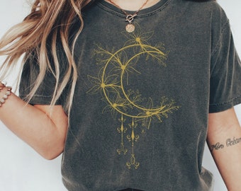 Op de afbeelding: Een donkergrijs T-shirt met een gouden bloemen-maanontwerp. De maan is omringd door bloemen en wijnstokken, en er hangen charmes aan de onderkant. Het shirt wordt gedragen door een persoon met lang bruin haar en een tatoeage op hun arm die "More Dreams" zegt.