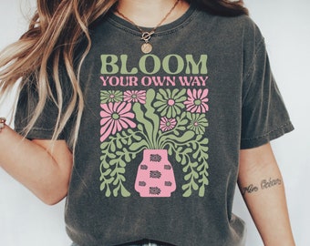 Puede incluir: Camiseta gris oscuro con el texto "BLOOM YOUR OWN WAY" sobre un diseño floral. El diseño presenta flores rosas y verdes en un jarrón rosa. La camiseta se combina con vaqueros azules y zapatillas blancas.