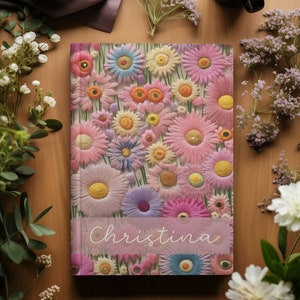 Pode incluir: Um caderno personalizado com um design floral com flores rosa, amarelas e azuis. O nome "Christina" est&aacute; impresso na capa.