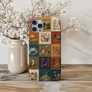 Puede incluir: Una funda de teléfono rectangular con una cuadrícula de parches estilo bordado. Cada cuadrado muestra una escena diferente, incluyendo un amanecer sobre colinas, una cabaña, un bote en un río, un búho, un ciervo, un zorro y setas.