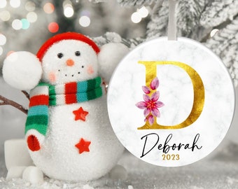 Può includere: Un ornamento natalizio in marmo bianco con una lettera D dorata e accenti floreali rosa. L'ornamento è personalizzato con il nome "Deborah" e l'anno "2023".