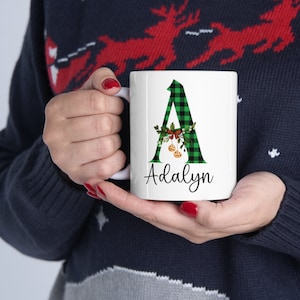 May include: White ceramic mug with a green and black buffalo plaid letter A with a Christmas wreath and ornaments. The mug has the name "Adalyn" written below the letter.