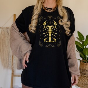 May include: Black t-shirt with a gold scorpion zodiac symbol and other astrological symbols. The scorpion is surrounded by a circle with stars and a crescent moon.