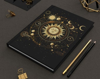 May include: A black notebook with a gold celestial design featuring a sun, moon, and stars. The notebook is closed and lying on a dark gray surface.