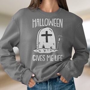 Puede incluir: Sudadera gris con un gráfico de una lápida con una cruz y una mano de esqueleto. El texto "Halloween gives me life" está impreso debajo del gráfico.