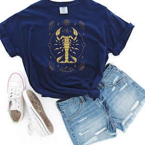 May include: A navy blue t-shirt with a gold scorpion graphic and astrological symbols. The scorpion is surrounded by a circle, stars, and pyramids.