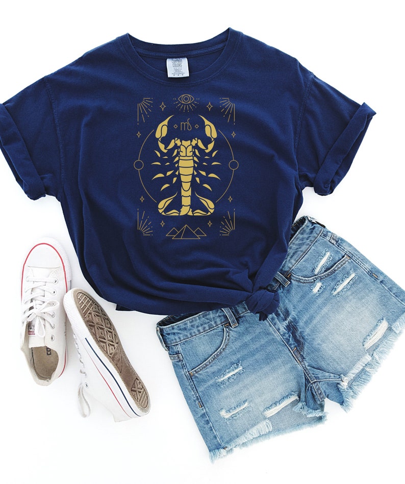 May include: A navy blue t-shirt with a gold scorpion graphic and astrological symbols. The scorpion is surrounded by a circle, stars, and pyramids.