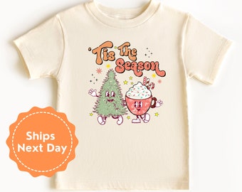 May include: A cream coloured t-shirt with a Christmas themed graphic. The graphic features a cartoon Christmas tree and a cartoon cup of hot chocolate with whipped cream and candy canes. The text "'Tis the Season" is written in a retro font above the graphic.