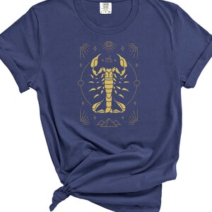 May include: A navy blue t-shirt with a gold scorpion graphic and astrological symbols. The scorpion is surrounded by a circle with stars and a pyramid at the bottom.