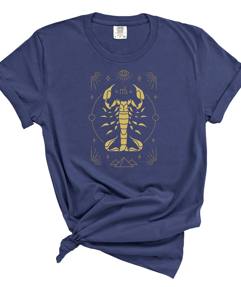 May include: A navy blue t-shirt with a gold scorpion graphic and astrological symbols. The scorpion is surrounded by a circle with stars and a pyramid at the bottom.