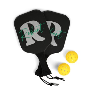 May include: Two black and green personalized pickleball paddles with the name "Robert" on one paddle and "Bert" on the other paddle. The paddles are in a black drawstring bag and two yellow pickleball balls are in the foreground.