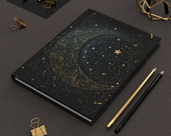 May include: A black notebook with a gold crescent moon and stars design. The notebook is lying on a dark surface with two pencils and gold paper clips.