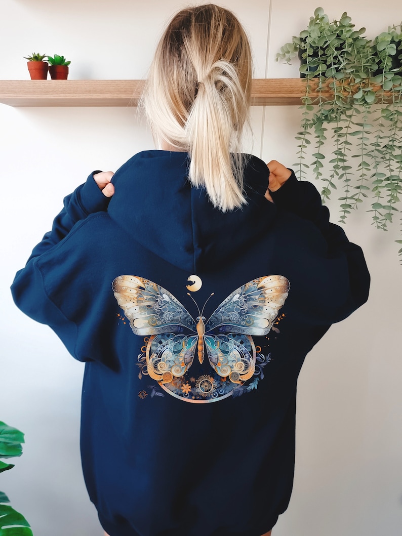 Pode incluir: Moletom azul marinho com um design detalhado de borboleta nas costas. A borboleta &eacute; em tons de dourado, azul e branco, com uma lua crescente acima. O moletom tem capuz com cord&atilde;o e mangas compridas.