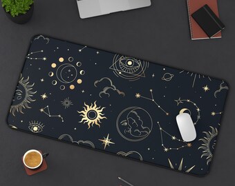 May include: A black rectangular desk mat with a celestial design featuring gold suns, moons, stars, and constellations. A white mouse rests on the mat, alongside a laptop, coffee cup, and stationery.