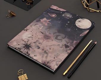 May include: A hardcover notebook featuring a dark celestial and floral print with a full moon, stars, and soft pink flowers. The notebook is accompanied by a gold-toned pencil, a black pencil, and several gold-toned metal binder clips.