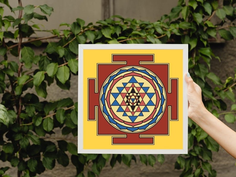 May include: A framed print of a Sri Yantra, a sacred geometric symbol in Hinduism, with a yellow background, red and blue accents, and a white frame.