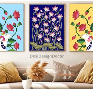 Op de afbeelding: Drie ingelijste schilderijen met een blauwe, gele en lichtblauwe achtergrond. Elk schilderij toont een witte koe met een bloemenmotief en roze bloemen.