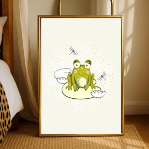 May include: A framed art print featuring a whimsical illustration of a green frog sitting on a lily pad. The frog has large eyes and is surrounded by dragonflies and a dotted background. The frame is gold.