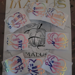 Könnte beinhalten: Neun schimmernde holografische Aufkleber mit herzförmigen Designs. Jeder Aufkleber zeigt einen anderen Spruch, darunter "to moon", "Orion", "keen hee", "seona", "xtion", "leedia", "raum", "n.woon" und "to moon".