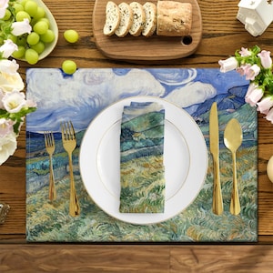 Può includere: Un runner da tavolo blu e verde con un dipinto di un paesaggio. Il runner è apparecchiato con un piatto bianco, posate d'oro e un tovagliolo piegato.