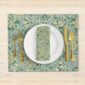 Pode incluir: Um tapete de mesa verde e branco com padrão floral, um prato branco, talheres dourados e uma toalha de mesa combinando.