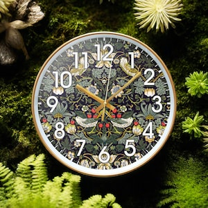 Könnte beinhalten: Eine weiße Uhr mit schwarzen römischen Ziffern und goldenen Zeigern. Das Zifferblatt der Uhr zeigt ein Blumenmuster mit Vögeln und Beeren. Die Uhr ist vor einem grünen moosbewachsenen Hintergrund platziert.